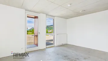 Heller Raum mit großen Fenstern, Betonboden und Zugang zu einem Balkon mit grüner Aussicht.