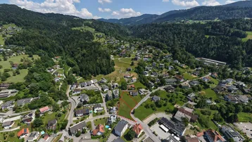 Luftaufnahme eines Baugrundstücks in einer grünen Wohngegend mit bewaldeten Hügeln im Hintergrund.