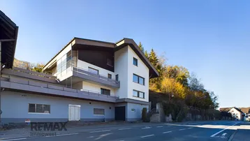 Modernes Gebäude mit mehreren Etagen, Balkonen und einer Straße im Vordergrund unter blauem Himmel.