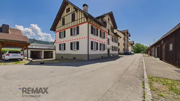 Traditionelles Mehrfamilienhaus mit Holzfassadendetails und umliegender Zufahrt unter blauem Himmel.