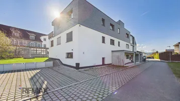 Modernes Mehrfamilienhaus mit weißer Fassade, Schieferdach und Außenparkplätzen unter strahlendem Himmel.