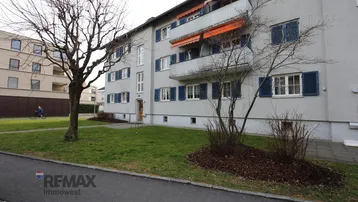 Mehrfamilienhaus mit gepflegter Grünfläche und blauen Fensterläden in städtischer Umgebung.