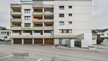 Mehrstöckiges Wohngebäude mit Balkonen, Garagen und Außenparkplätzen vor bewölktem Himmel.