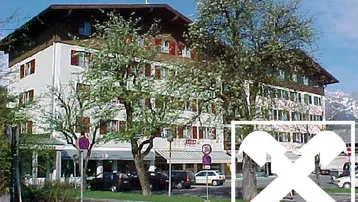 Mehrstöckiges Gebäude mit traditioneller Architektur, roten Fensterläden und Balkonen, umgeben von Bäumen und einer Straße, mit Bergen im Hintergrund.