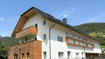 Modernes Mehrfamilienhaus mit Holzfassade und Balkonen in ländlicher Umgebung und Bergblick.