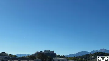 Panoramablick über die Stadt Salzburg mit der Festung Hohensalzburg und den umliegenden Bergen unter blauem Himmel.