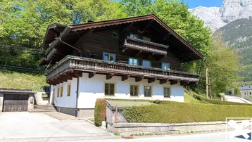 Traditionelles Haus mit Holzbalkonen und weißer Fassade, umgeben von grünen Bäumen und Bergen.