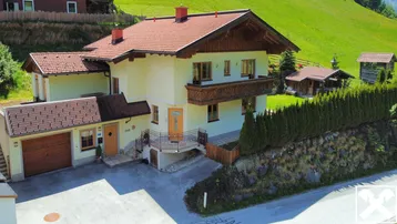 Traditionelles Haus im alpinen Stil mit rotem Ziegeldach und Balkon, umgeben von grünen Hügeln.