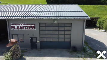 Ansicht der Gewerbehalle mit großem Rolltor und Firmenbeschriftung vor einer grünen Hügellandschaft.