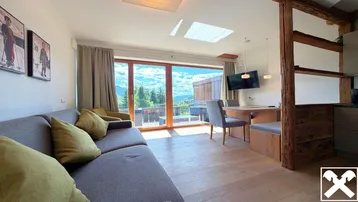 Heller Wohn- und Essbereich mit Holzboden, freiliegenden Holzbalken und Zugang zu einem Balkon mit Bergblick.
