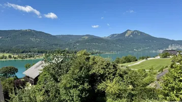 Panoramablick auf einen türkisfarbenen See, umgeben von grünen Bergen und üppiger Vegetation unter blauem Himmel.