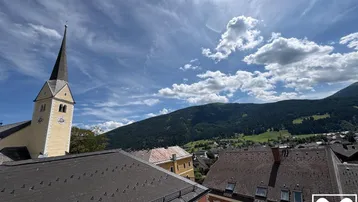 Blick über Dächer auf eine Kirche, ein Dorf und grüne Berge unter blauem Himmel.