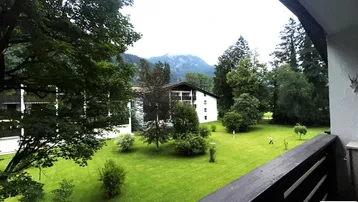 Blick vom Balkon auf eine grüne Wiese mit Bäumen und Bergen im Hintergrund.