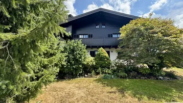 Traditionelles Holzhaus mit dunkler Fassade, umgeben von üppigem Grün und Bäumen.