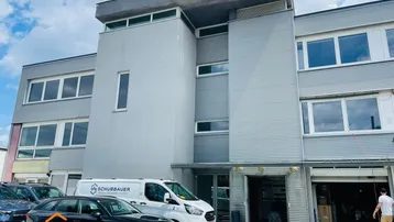 Modernes Geschäftsgebäude mit mehreren Etagen, großen Fenstern und Parkplätzen davor.