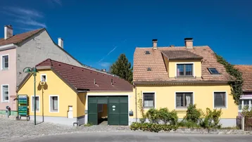 Gelbes Wohnhaus mit rotem Ziegeldach und angrenzendem Garagengebäude an einer ruhigen Straße.