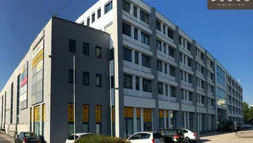 Modernes Bürogebäude mit mehreren Etagen und großen Fenstern, umgeben von Parkplätzen unter blauem Himmel.