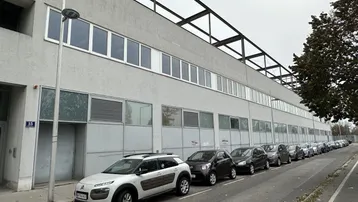 Lange Fassade eines Industriegebäudes mit parkenden Autos entlang der Straße unter bewölktem Himmel.