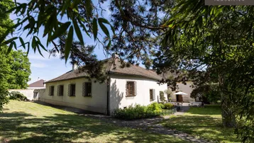 Historisches Gebäude umgeben von einem weitläufigen Garten mit altem Baumbestand unter blauem Himmel.