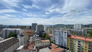 Weitläufiger Blick über die Stadt mit Wohngebäuden und Bergen im Hintergrund unter blauem Himmel.
