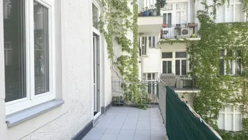 Heller Balkon mit Fliesenboden und Blick auf begrünte Fassaden im Innenhof eines Altbaus.