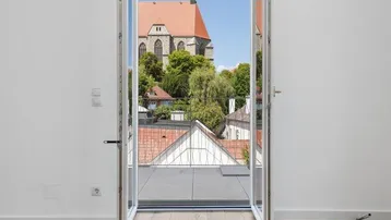 Große Fenstertür mit Blick auf die Dächer und eine historische Kirche, heller Holzboden.