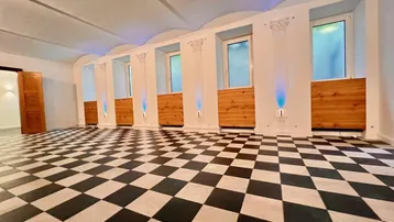 Geräumiger, loftartiger Raum mit auffälligem schwarz-weißem Schachbrettmuster-Boden, hohen Gewölbedecken und dekorativen Säulen.