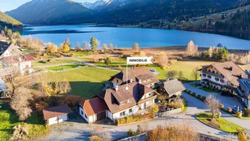 Luftaufnahme des Anwesens mit umliegenden Häusern, See und Bergpanorama im Hintergrund.