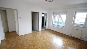 Helles Zimmer mit Parkettboden, weißen Wänden und Zugang zu weiteren Räumen.