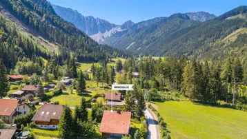Luftaufnahme eines Hauses in einem malerischen Tal, umgeben von Wäldern und hohen Bergen.