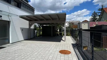Gepflasterter Außenbereich mit Carport und Zufahrt zum Gebäude unter blauem Himmel.