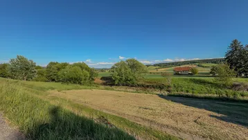 Ländliche Landschaft mit weiten Feldern und einem Haus in der Ferne unter blauem Himmel.