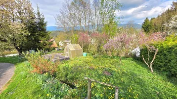 Hanglage im Garten mit einem Gewächshaus, blühenden Bäumen und einem weiten Blick über die ländliche Umgebung.