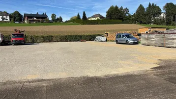 Großer Schotterparkplatz mit einem LKW und einem Van, umgeben von Feldern und Wohnhäusern im Hintergrund.
