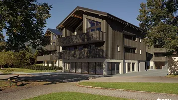 Neubau im alpinen Stil mit dunkler Holzfassade und großen Fenstern, umgeben von Grünflächen.