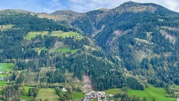 Weitläufiger Blick über ein grünes Tal mit bewaldeten Bergen und kleinen Siedlungen unter blauem Himmel.