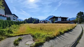 Unbebautes Grundstück mit Wiese und Blick auf die umliegende Berglandschaft unter blauem Himmel.