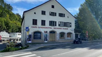 Ansicht der Gebäudefassade mit Geschäften im Erdgeschoss und Bergen im Hintergrund unter blauem Himmel.