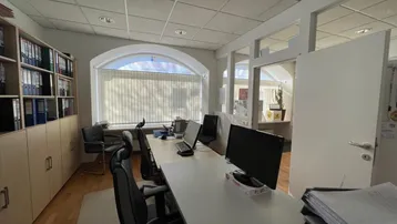 Helles Büro mit mehreren Arbeitsplätzen, Holzregalen und einem großen Bogenfenster mit Jalousien.