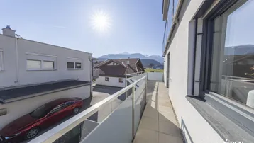 Geräumiger Balkon mit Glasgeländer, Fliesenboden und weitem Blick auf die Berge.