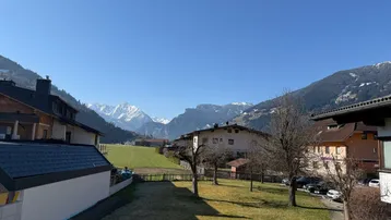 Weitläufiger Blick auf grüne Wiesen und schneebedeckte Berge unter einem klaren blauen Himmel.