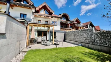 Reihenhaus mit gepflegtem Garten, Terrasse und Blick auf die umliegende Berglandschaft unter blauem Himmel.