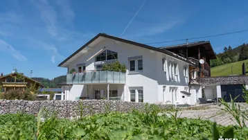 Modernes Wohnhaus mit weißer Fassade, Balkon und Blick auf die grüne Umgebung unter blauem Himmel.