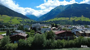 Panoramablick auf eine Stadt im Tal, umgeben von grünen Bergen unter blauem Himmel.