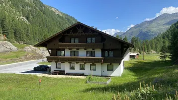 Traditionelles Alpenhaus mit Holzbalkonen und weißer Fassade, umgeben von grünen Wiesen und Bergen.