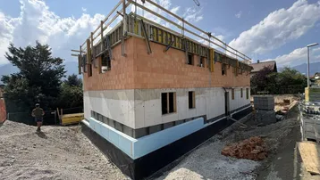 Rohbau eines neuen Gebäudes mit Ziegel- und Betonwänden, umgeben von Baustellengelände und Bergen.
