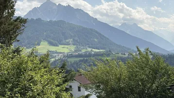 Malerischer Blick auf grüne Hügel, ein kleines Dorf und majestätische Berge unter einem teilweise bewölkten Himmel.