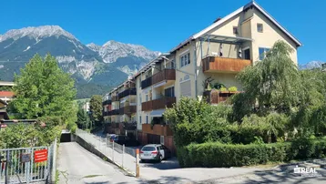 Mehrfamilienhaus mit Balkonen und Zufahrt, umgeben von Bäumen und mit Bergblick.