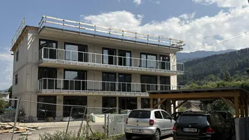 Modernes Mehrfamilienhaus im Bau mit Balkonen und Carport, umgeben von Berglandschaft.