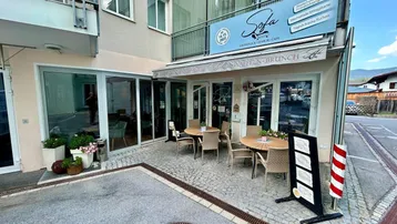 Außenbereich eines Cafés mit Tischen und Stühlen auf gepflasterter Terrasse unter einer Markise.